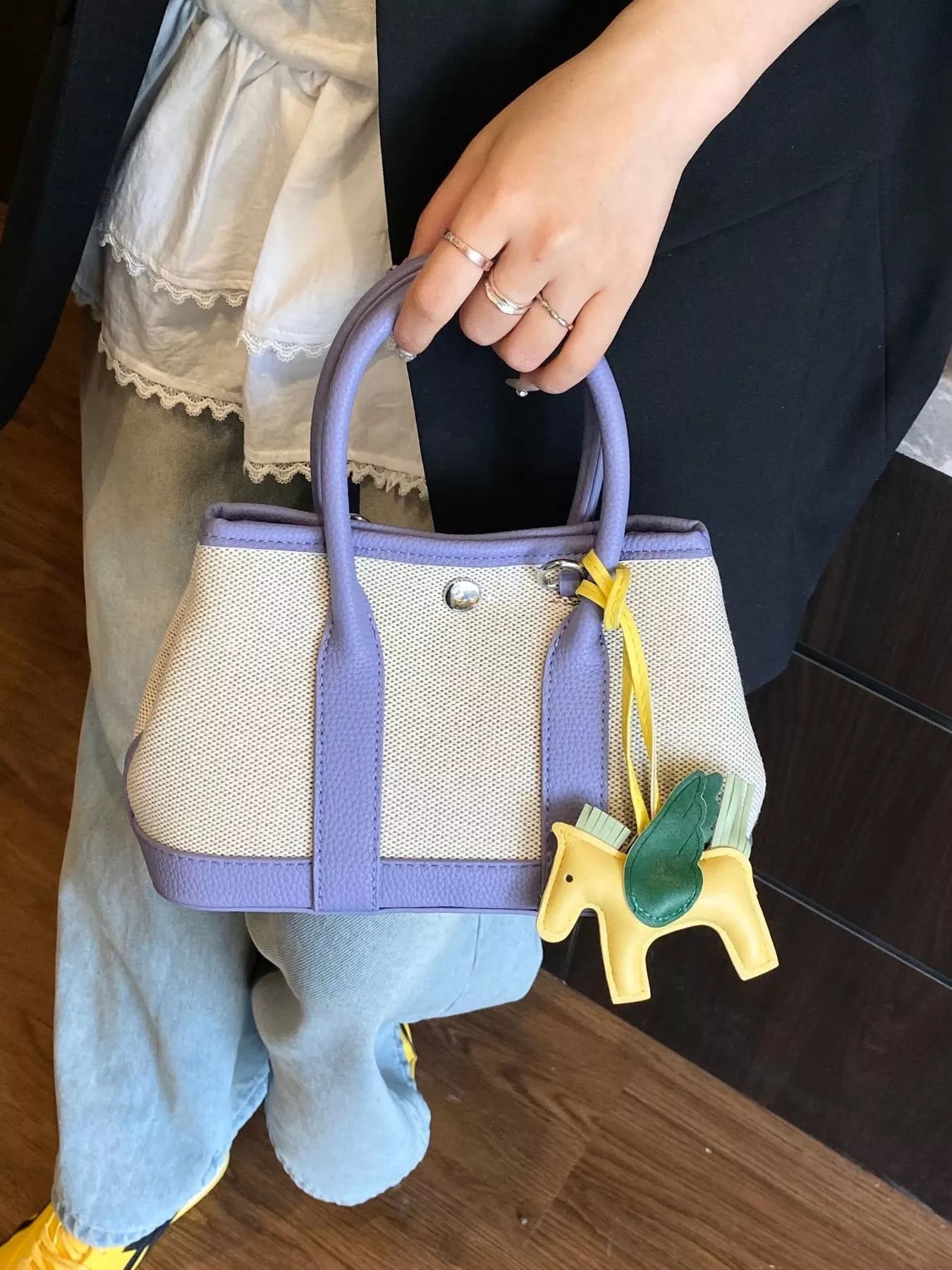 Kleine Handtasche aus beigem Stoff mit lilafarbenen Griffen und Rand, verziert mit einem gelben Pferdeanhänger
