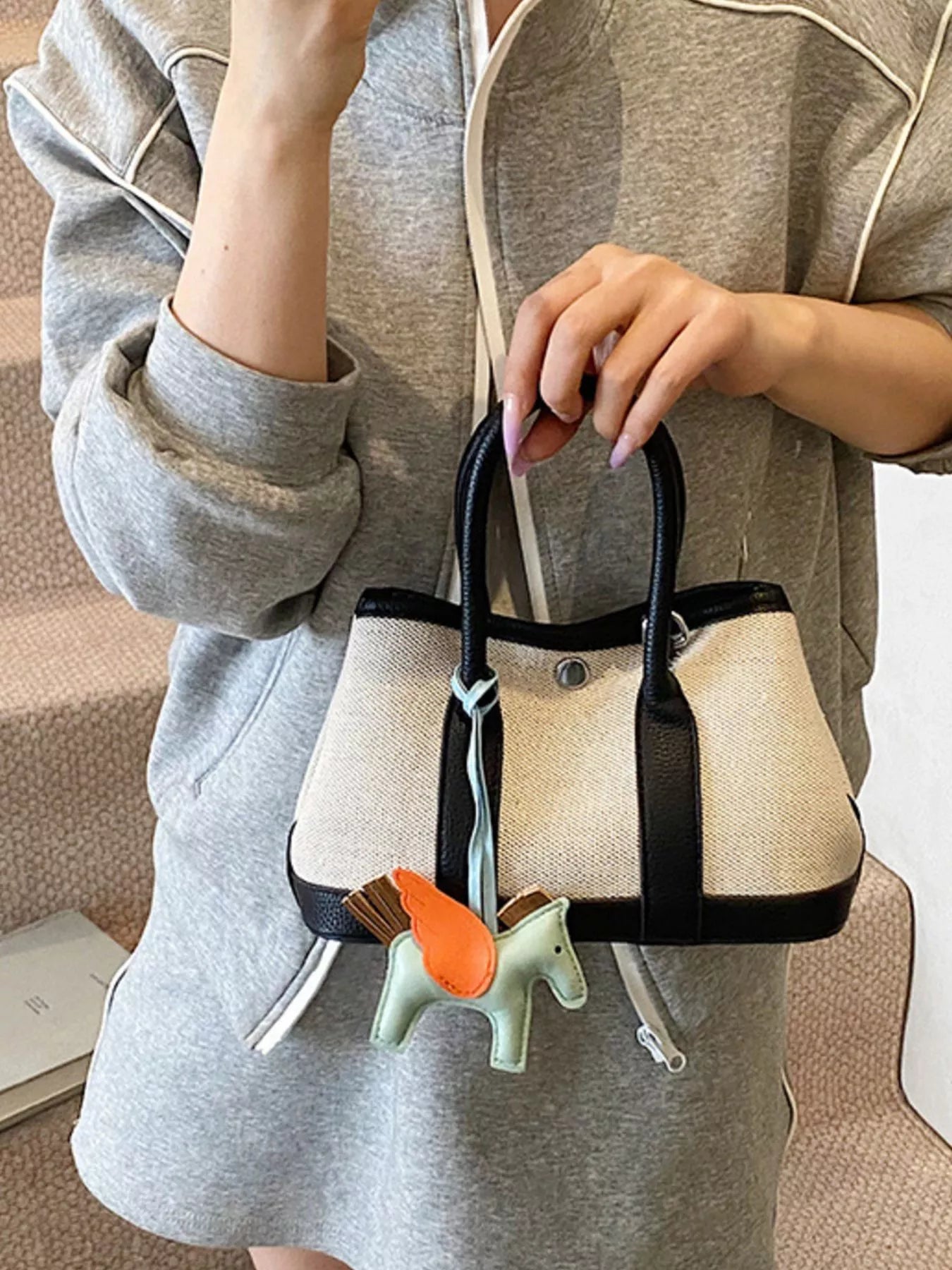 Kleine Handtasche aus beigem Stoff mit schwarzen Griffen und schwarzem Rand, verziert mit einem dekorativen Pferdeanhänger