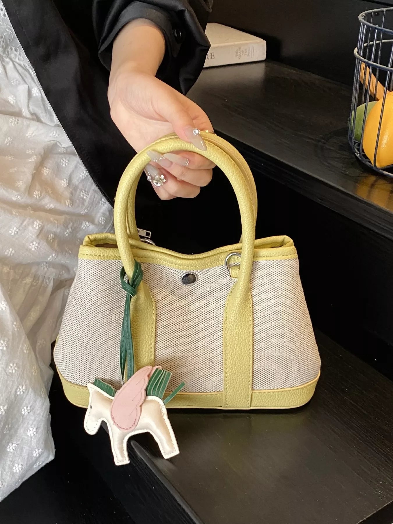 Kleine Handtasche aus beigem Stoff mit gelbgrünen Griffen und Rand, verziert mit einem dekorativen Pferdeanhänger.