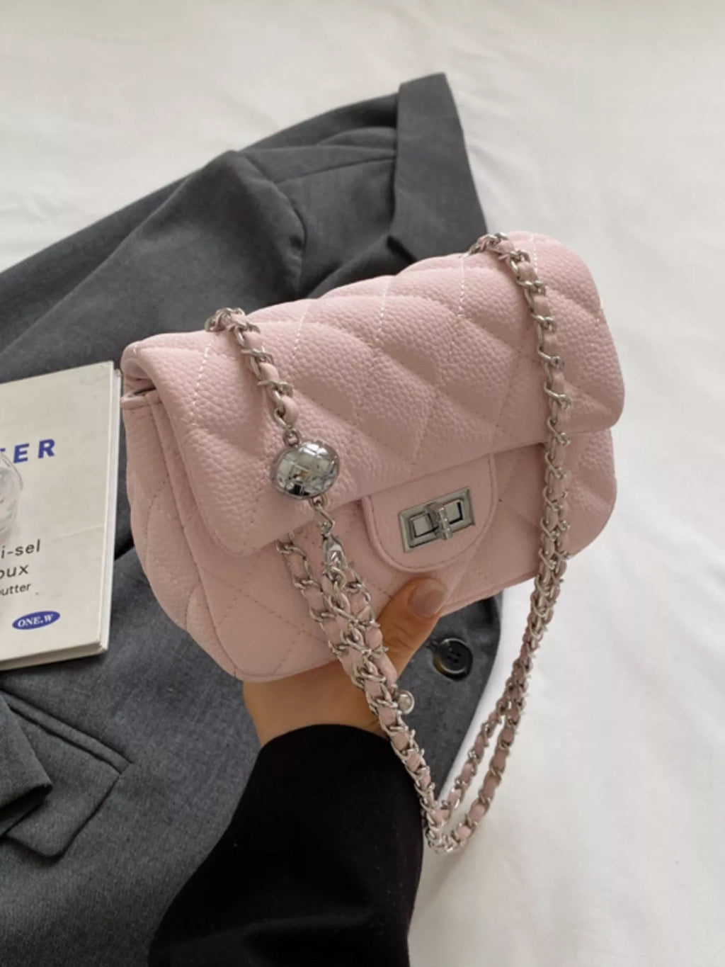 Lyra - Crossbody Tasche in zartem Rosa mit Ketten-Elementen auf einem grauen Hintergrund.
