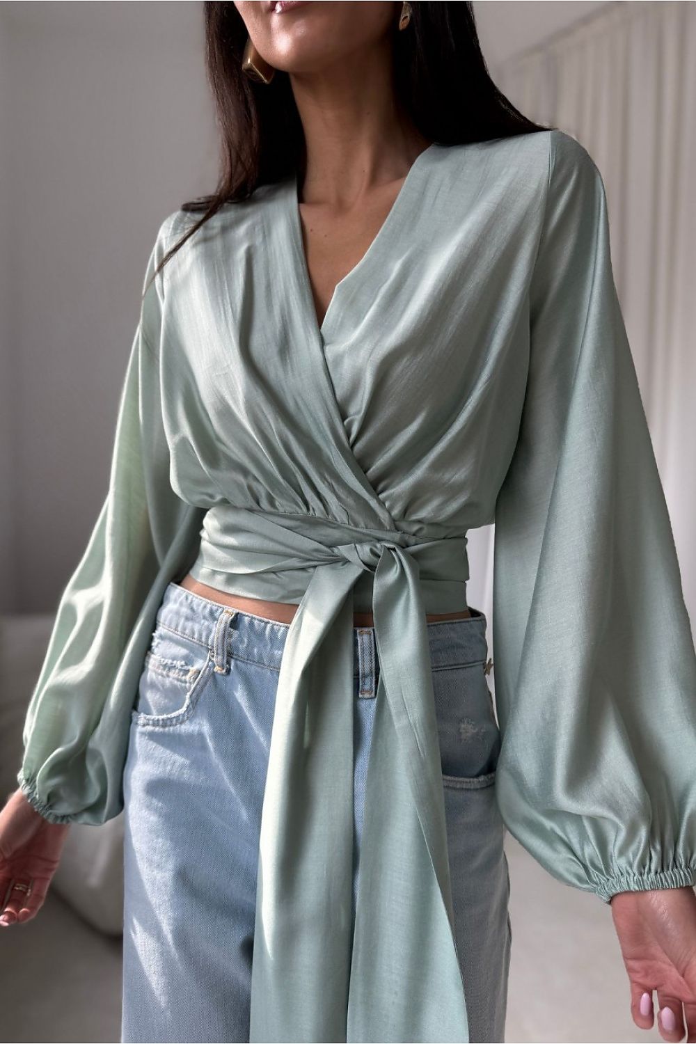 Trendige Kurzbluse in Salbeigrün (Sage Green) im Wickellook, kombiniert mit Ripped Jeans und goldenen Ohrringen für einen Casual-Chic-Look.